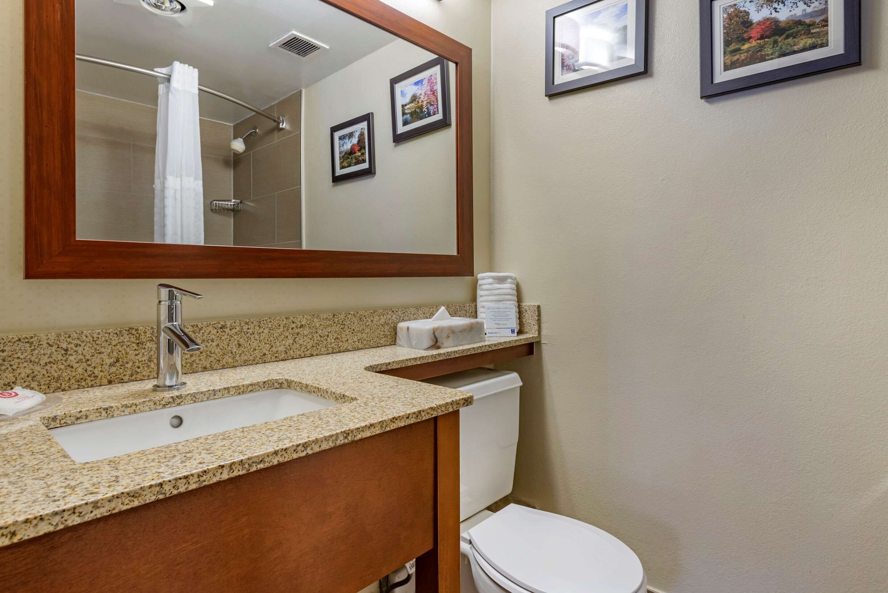 A bathroom with a granite countertop sink, a mirror, a toilet, and framed pictures on the wall.