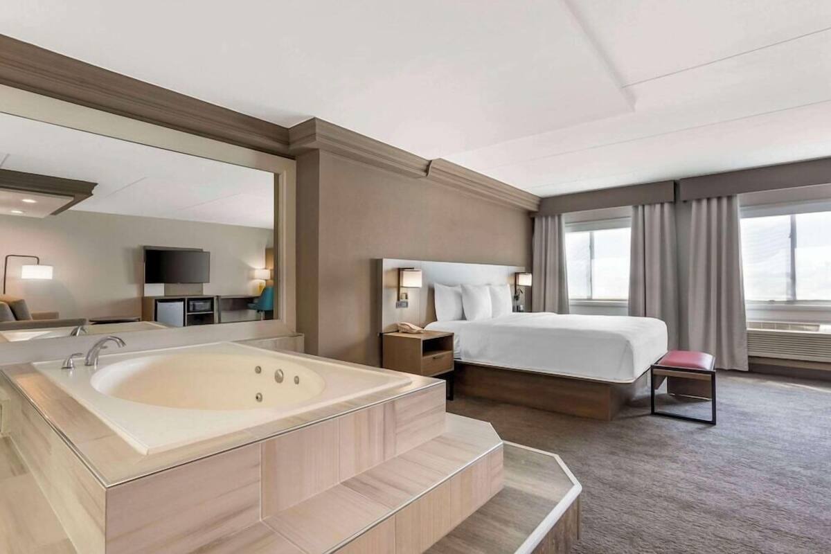 Une chambre d’hôtel avec une grande baignoire, un lit, une table de chevet, une télévision et une fenêtre avec des rideaux.