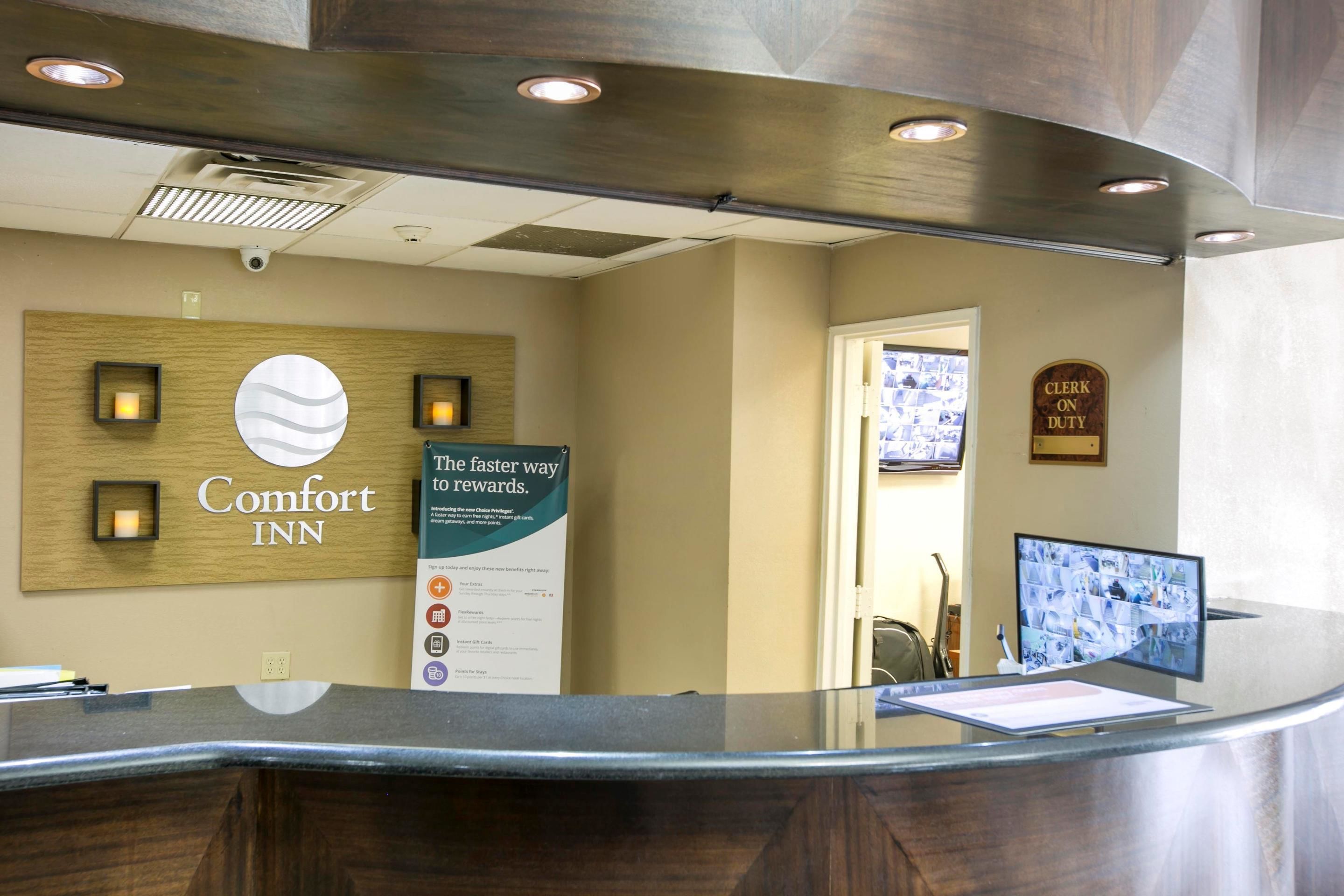 Un comptoir d’accueil dans un hôtel Comfort Inn, avec un panneau indiquant un moyen plus rapide d’obtenir des récompenses, un écran affichant des images de vidéosurveillance et un panneau indiquant la présence d’un réceptionniste.