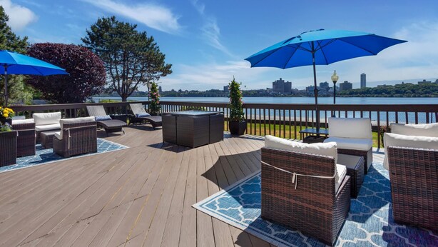 A deck with outdoor furniture and umbrellas, overlooking a body of water and city skyline.