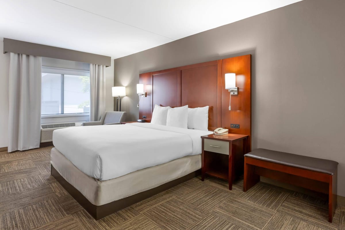 Une chambre d’hôtel avec un grand lit, des tables de chevet, un bureau, une chaise, un banc et une fenêtre avec des rideaux.