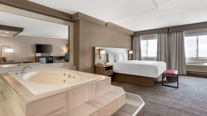 A hotel room with a large bathtub, a bed, a bedside table with a lamp, and a TV.