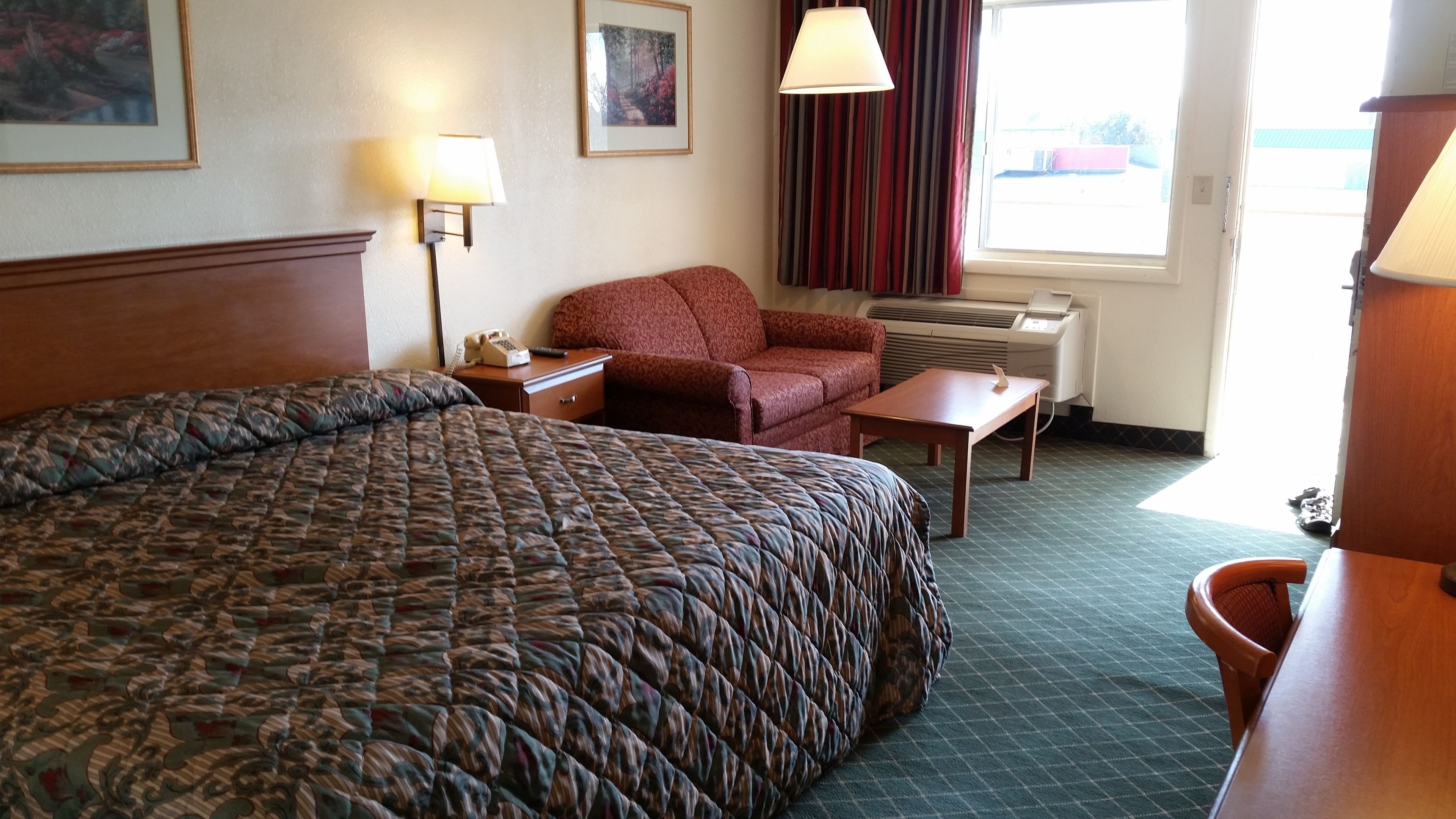 Habitación de hotel con una cama grande, una silla, una mesita, una lámpara, un cuadro en la pared y una ventana con cortinas.