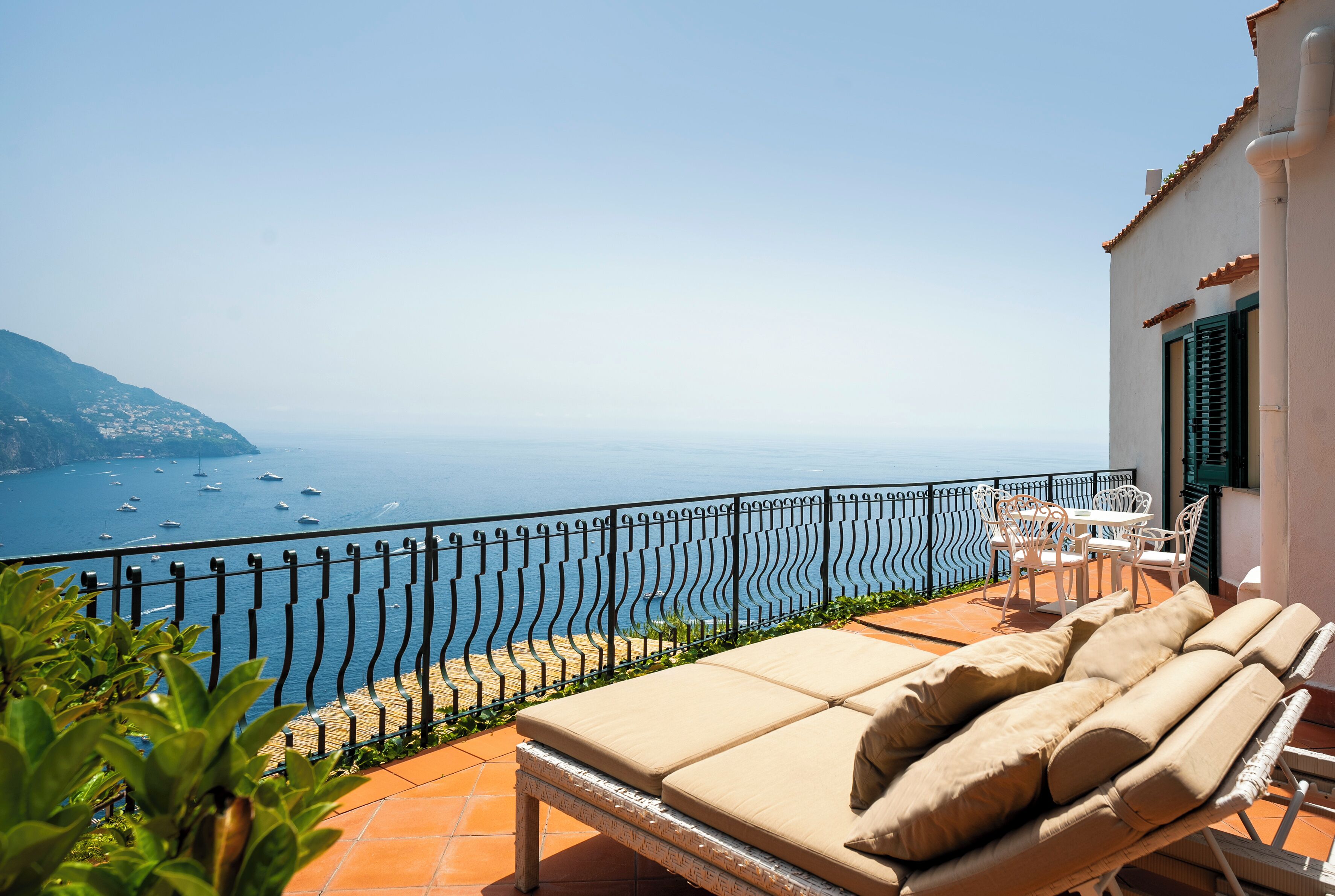 A balcony with a view of the sea, a white building, and outdoor furniture.