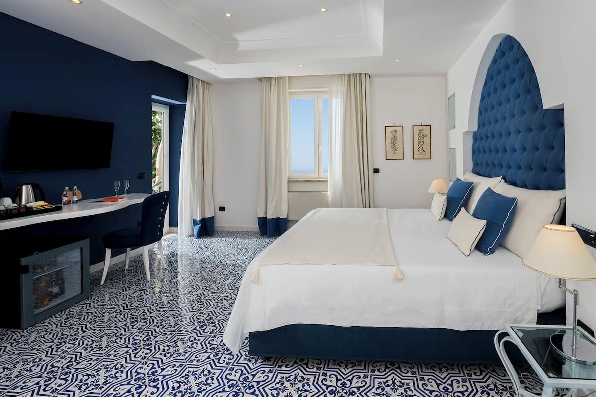 A hotel room with a large bed, a desk with a chair, a TV mounted on the wall, and patterned floor tiles.