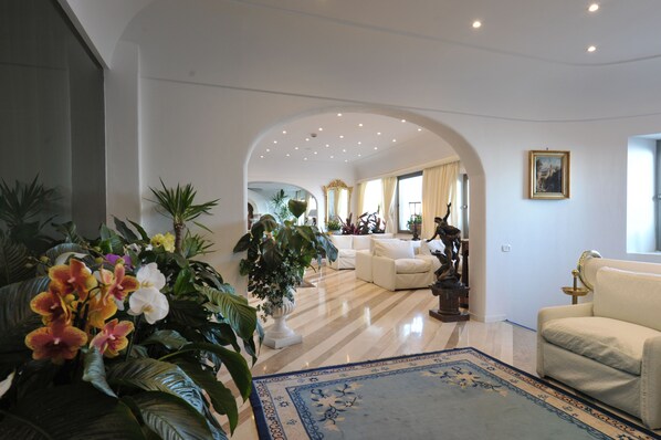 A spacious living room with a large white sofa, a coffee table, and a potted plant. There is a large window with curtains, a chandelier, and a framed picture on the wall.