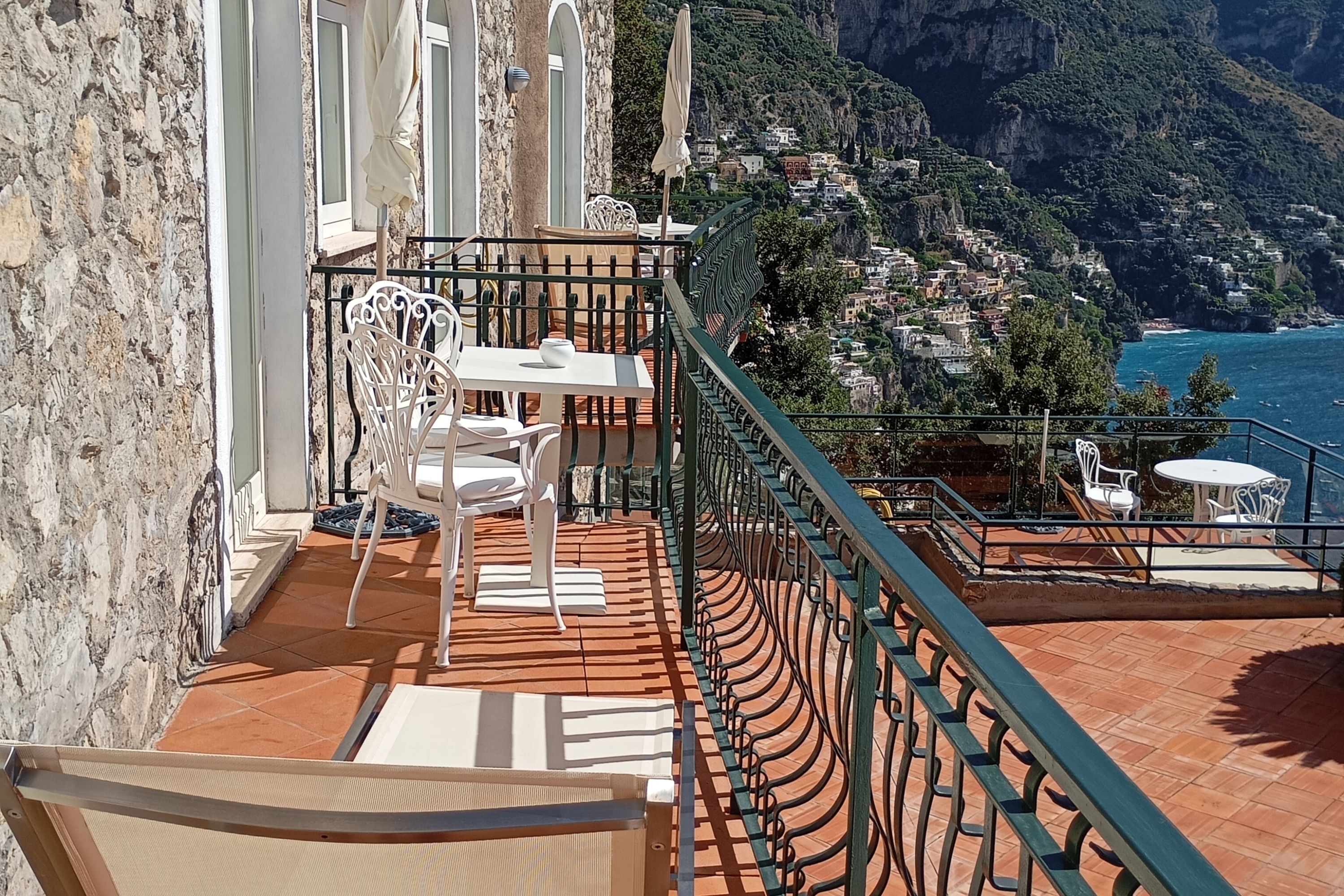Ein Balkon mit weißen Stühlen und einem Tisch, mit Blick auf eine Küstenstadt mit terrassenförmig angeordneten Häusern und Klippen.