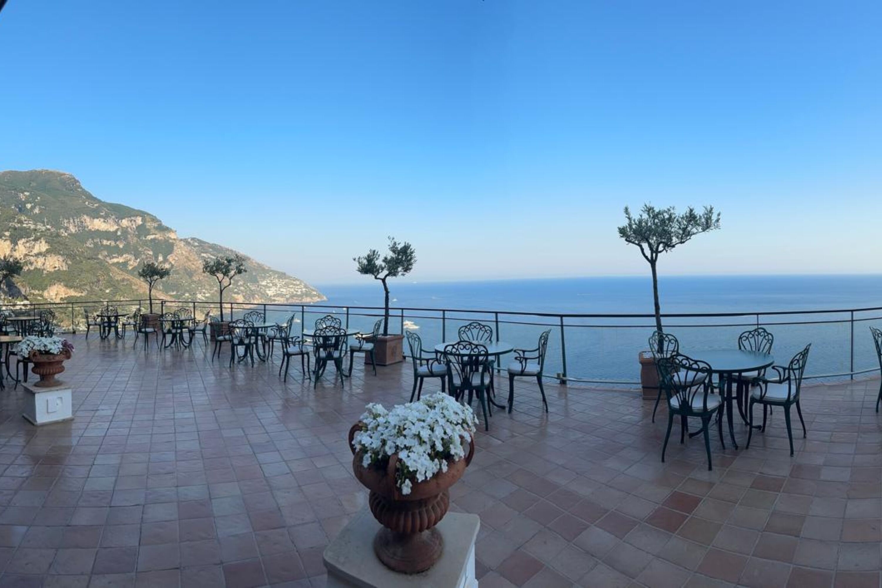 An outdoor terrace with tables and chairs, overlooking a body of water and a mountainous landscape.