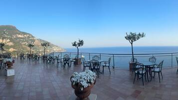 An outdoor terrace with tables and chairs, overlooking a body of water and a mountainous landscape.