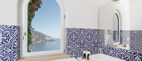 A bathroom with a view of the sea, featuring a white sink, a mirror, and blue patterned tiles.
