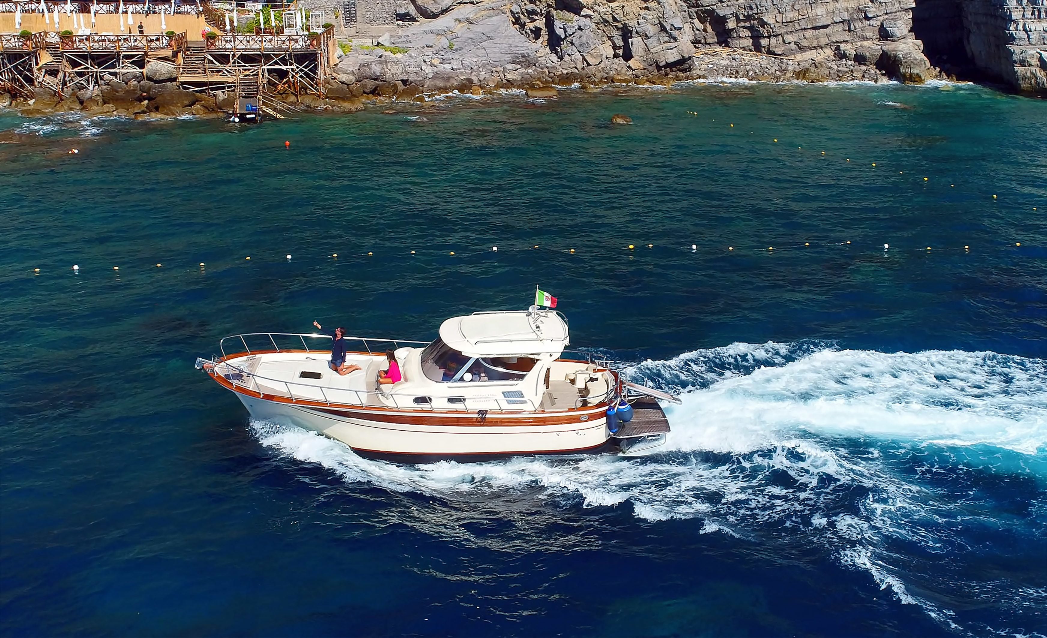 A boat with a flag, moving through clear water near rocky cliffs.