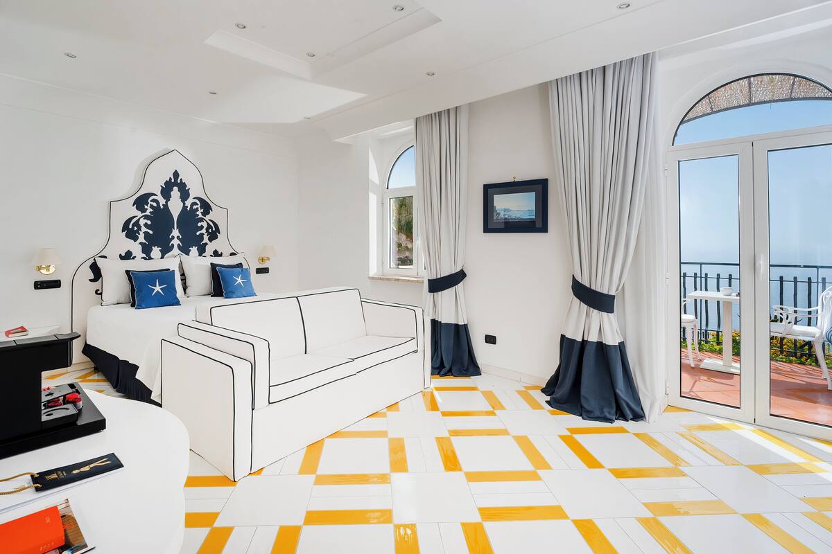 A modern bedroom with a white sofa, a large bed with a decorative headboard, and a balcony with a view.