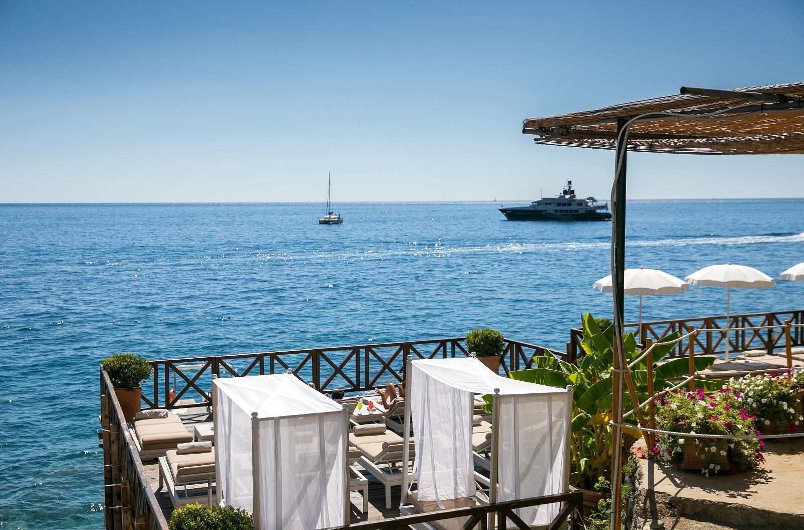 A seaside terrace with lounge chairs and umbrellas, overlooking the sea with boats in the distance.