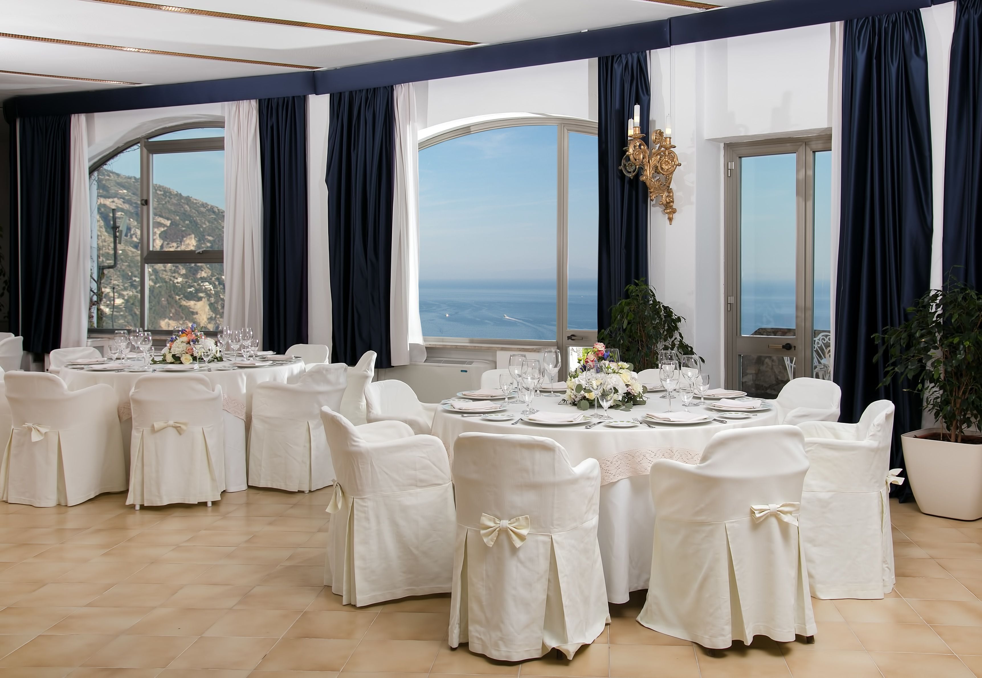 A banquet hall with round tables set for a formal event, large windows with ocean views, and elegant decor.