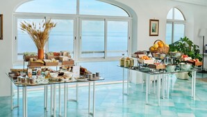 A spacious room with large windows overlooking the sea, featuring a buffet table with various food items and a decorative vase. - Hotel Le Agavi (Positano)