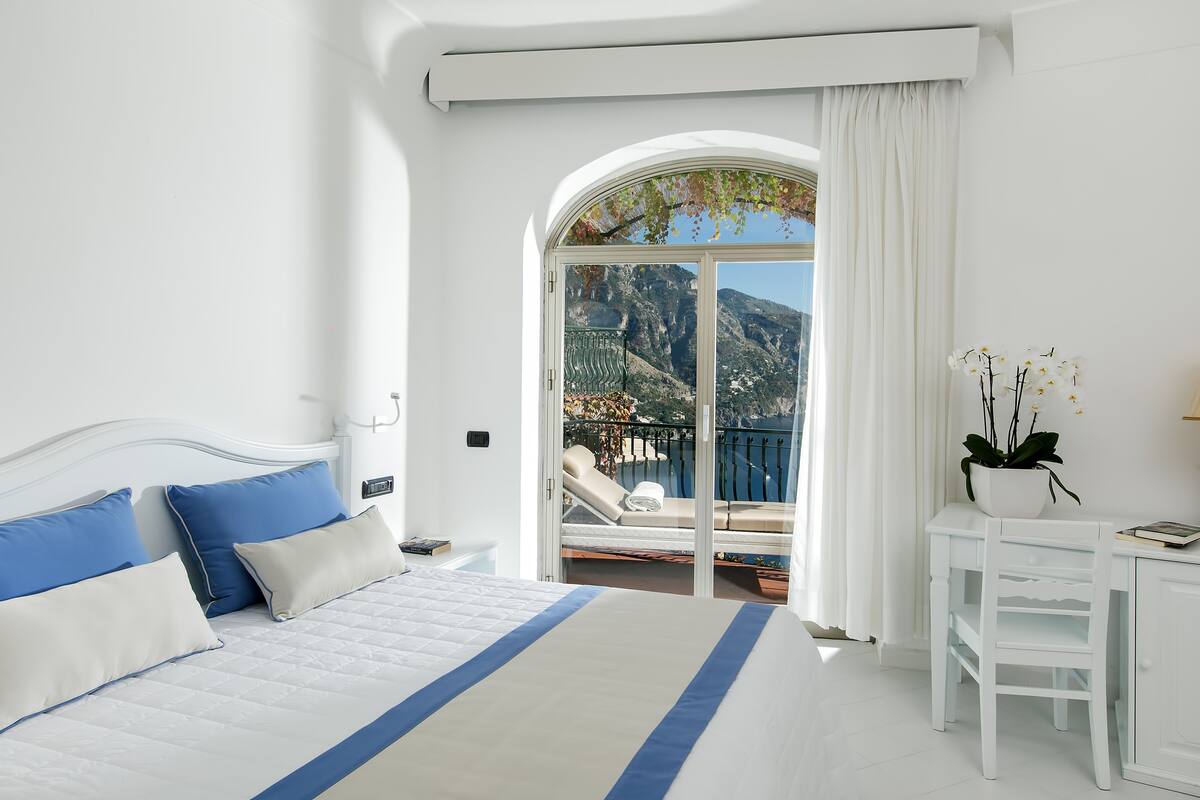 A bedroom with a bed, a nightstand, a chair, a potted plant, and a view of a balcony with a chair and a table.
