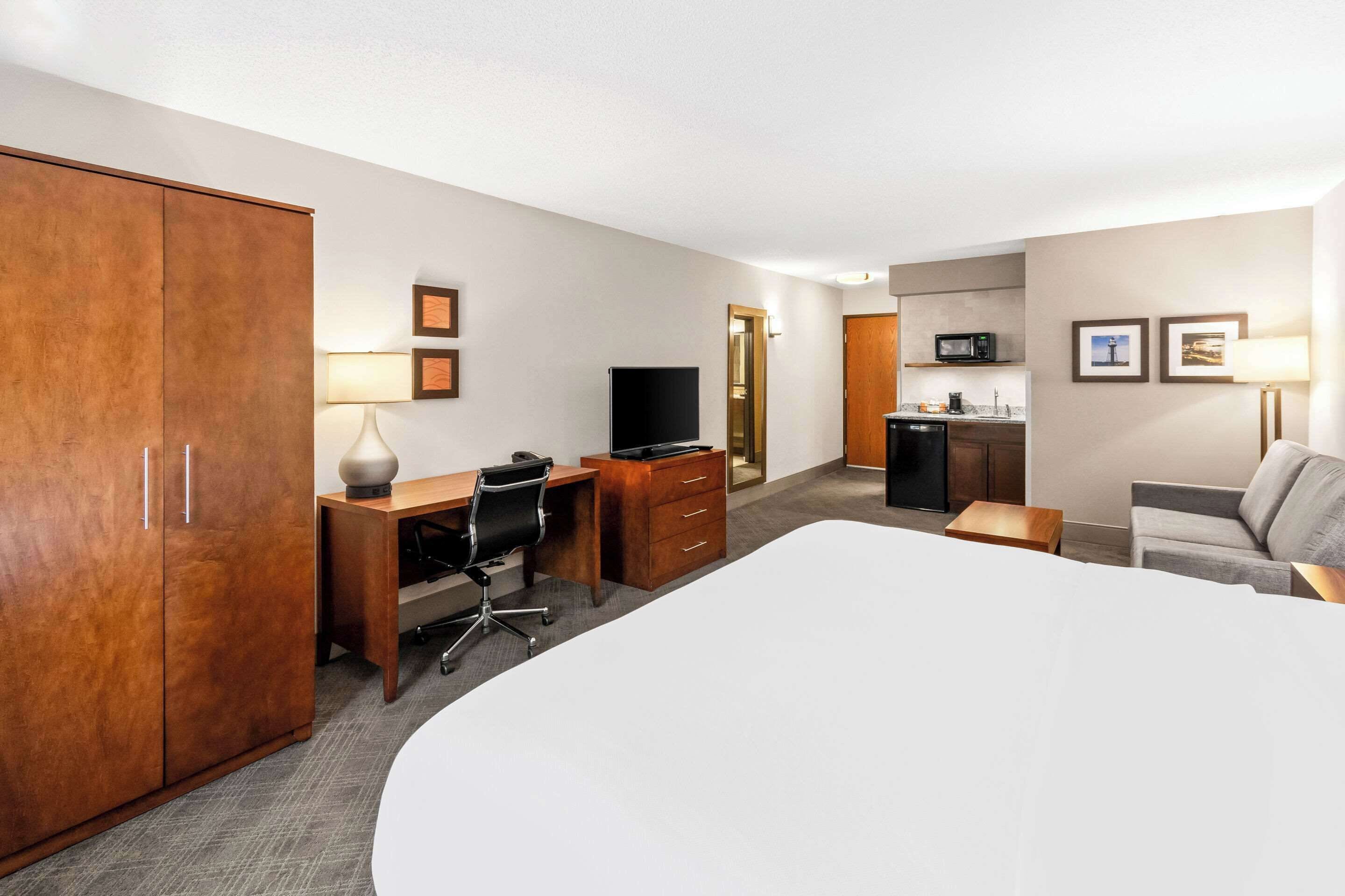 Ein Hotelzimmer mit einem großen Bett, einem Schreibtisch mit Stuhl, einem Fernseher, einem Kleiderschrank, einer Küchenzeile und einem Sofa.