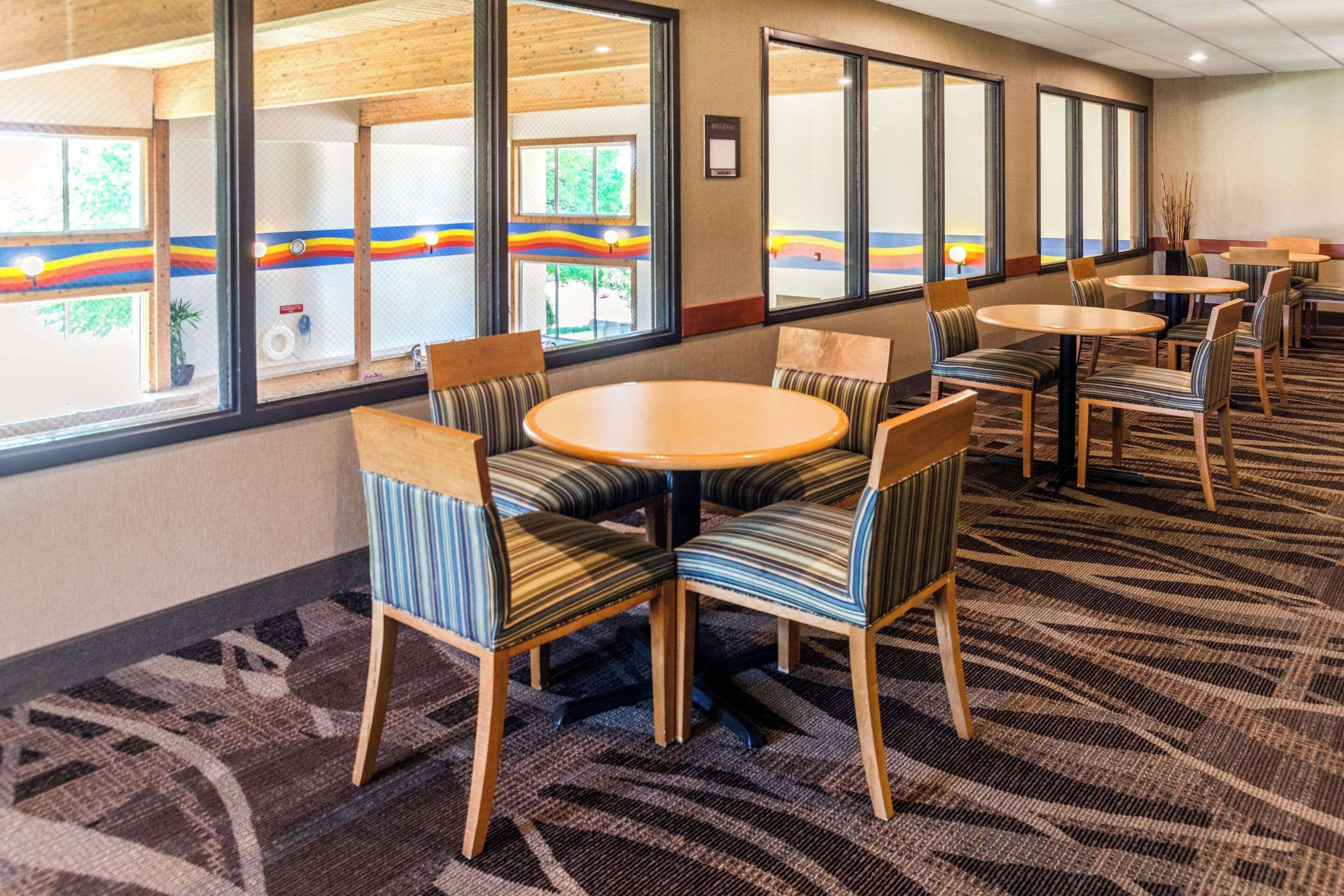 Un hall d’hôtel avec des tables rondes et des chaises à rayures, de grandes fenêtres offrant une vue dégagée et un tapis à motifs.