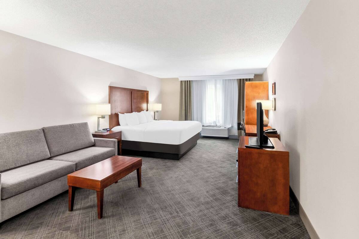 A hotel room with a bed, a sofa, a wooden coffee table, a desk with a lamp, and a television.