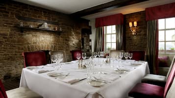 Un comedor con pared de piedra, una mesa preparada para seis personas con sillas tapizadas en rojo y una ventana con cortinas.