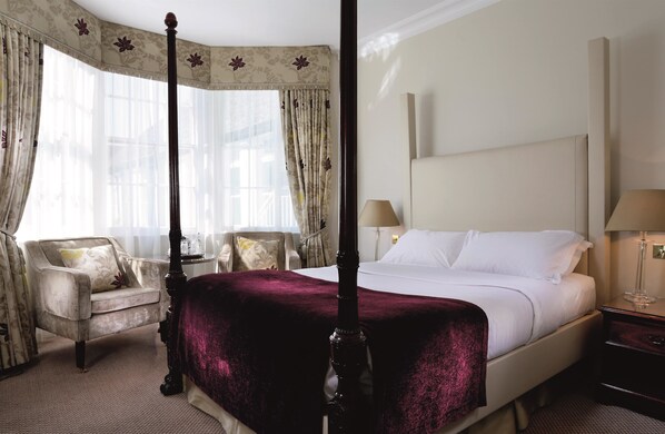 Una cama con dosel con un edredón rojo oscuro, un sillón con cojín floral, una mesita de noche con lámpara y una ventana con cortinas traslúcidas.