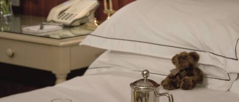 Una cama bien hecha con un peluche, una cafetera, una taza de café, un vaso de jugo de naranja y un periódico sobre una mesita de noche.