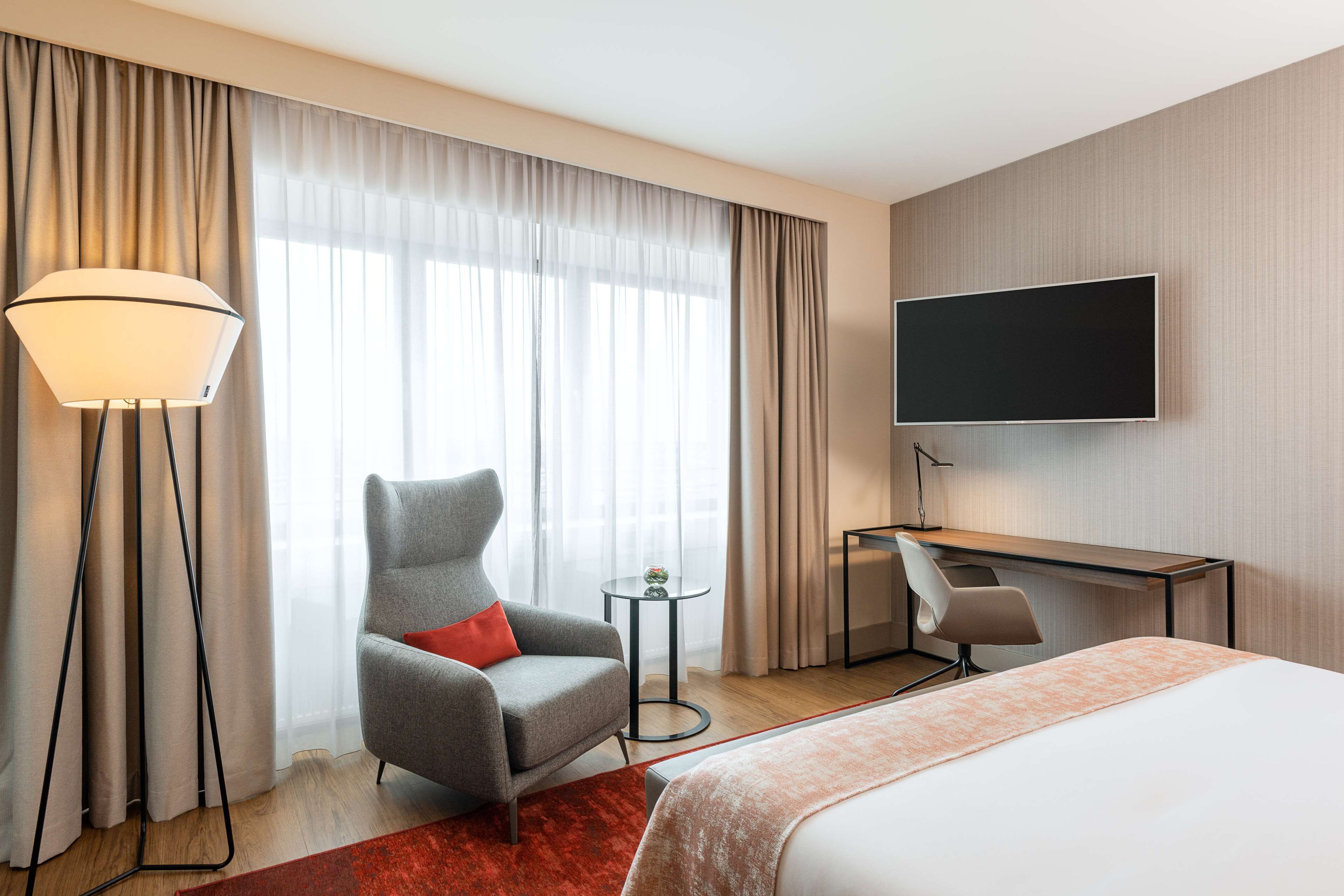 A hotel room with a bed, a desk, a chair, a TV, and a floor lamp.