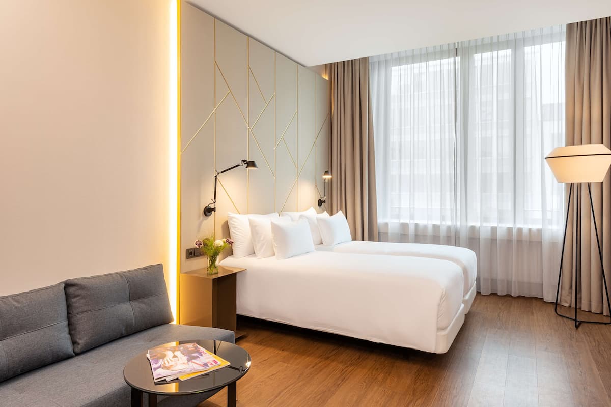 A hotel room with a double bed, a sofa, a small table with a magazine, and a lamp.