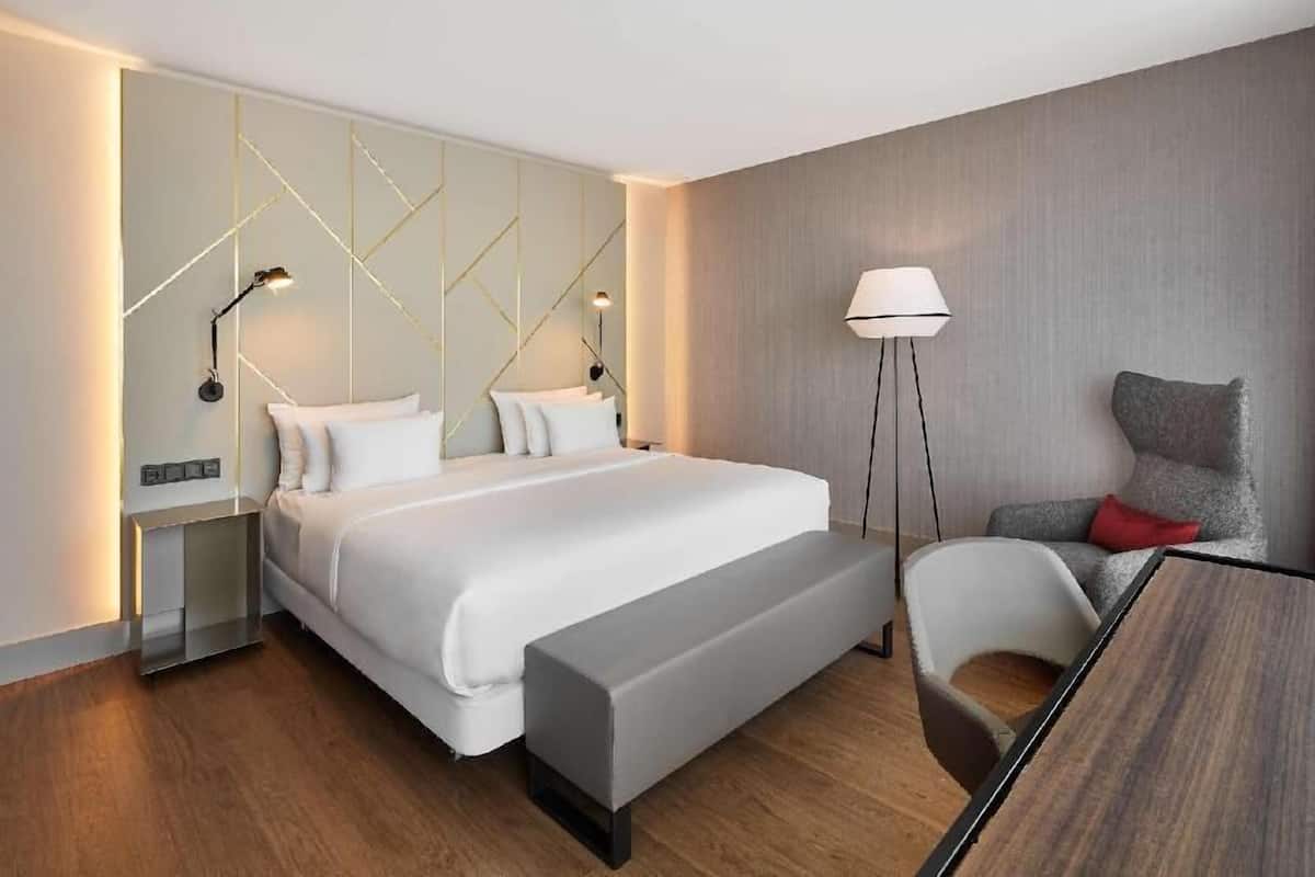 A modern hotel room with a large bed, bedside tables, a desk, a chair, and a floor lamp.