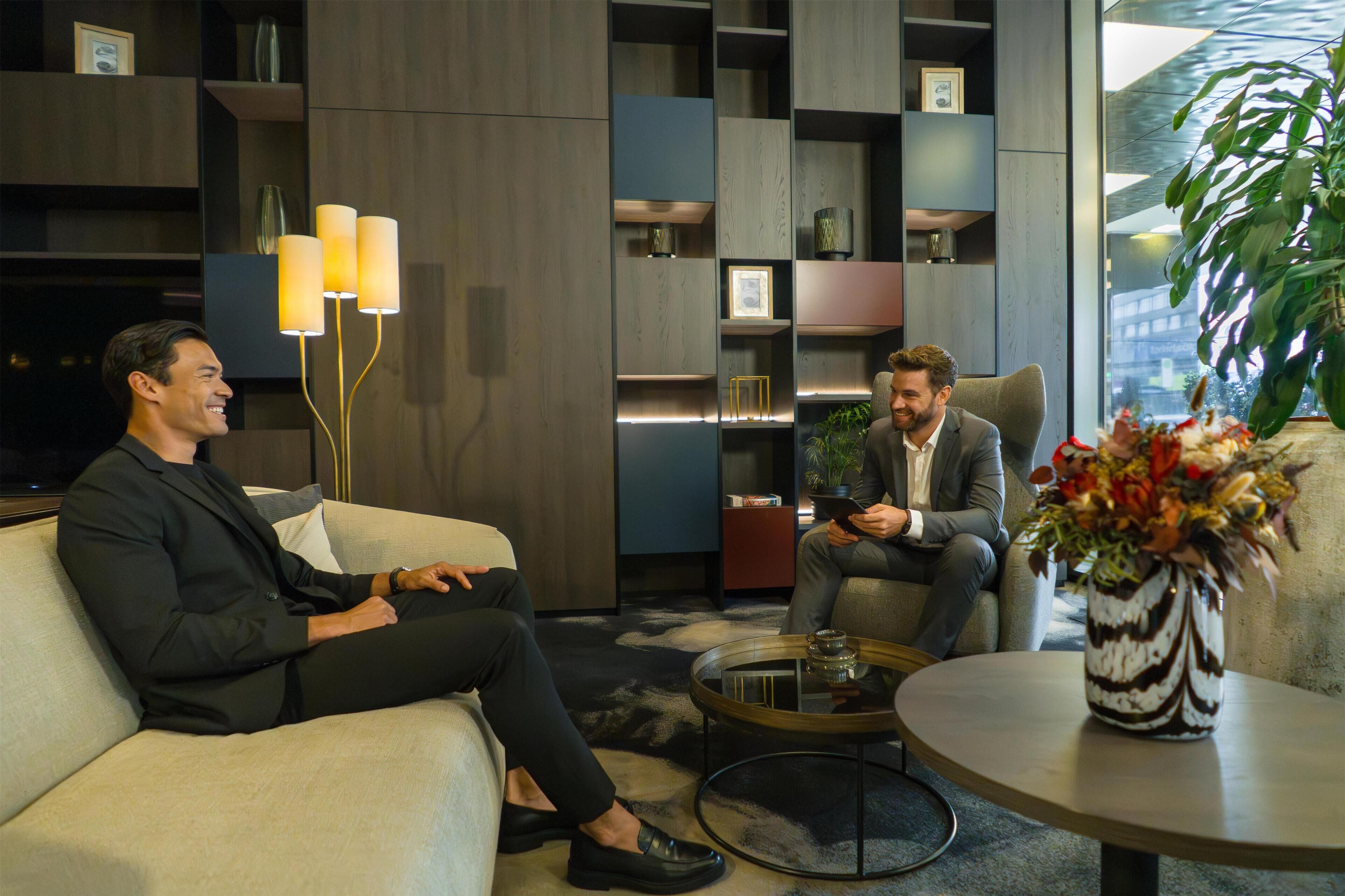 Dos hombres sentados en un sofá en una habitación moderna con estanterías empotradas y una mesa de centro.