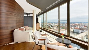 Un interior moderno con una pared curva de madera, un sofá, sillas y vistas a un paisaje urbano a través de amplios ventanales.