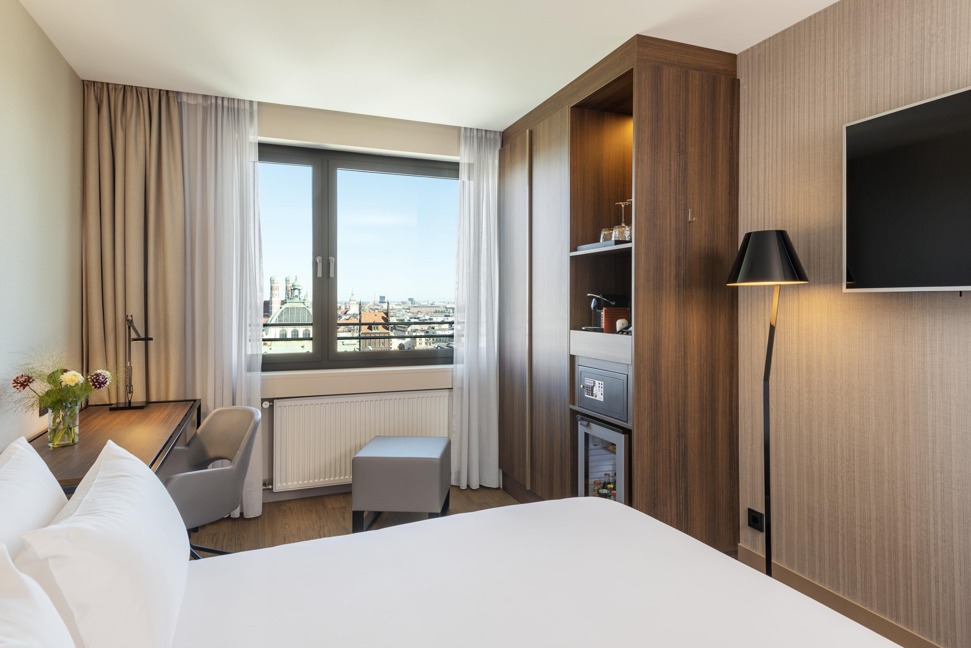 Une chambre d’hôtel comprenant un lit, un bureau, une chaise, une petite table avec un vase de fleurs, une télévision et une vue sur le paysage urbain.