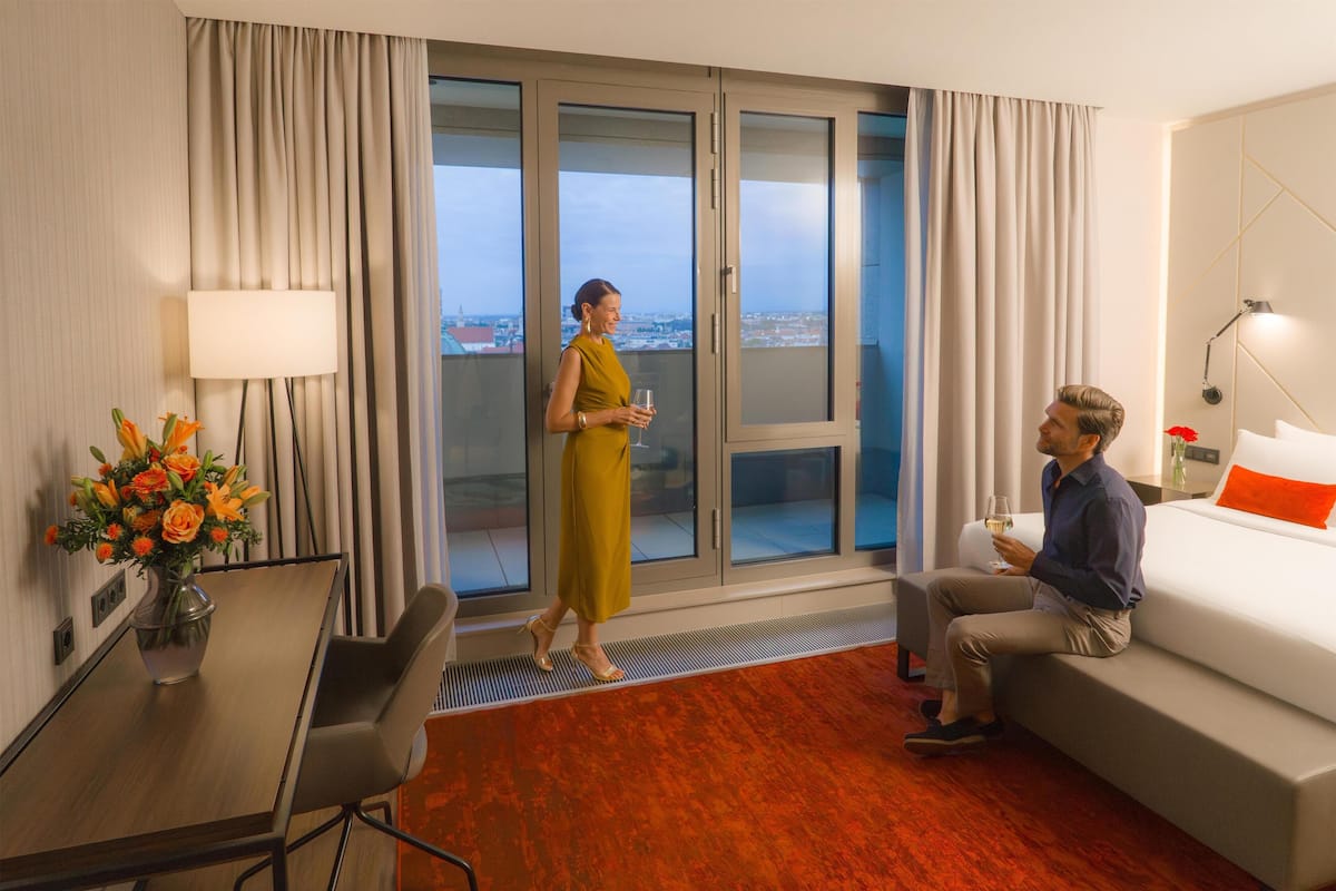 A hotel room with a balcony, a dining table with chairs, a sofa, and a bed with an orange pillow.