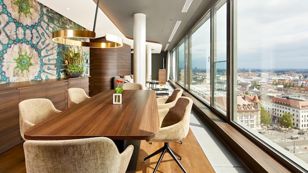Una sala de conferencias moderna con una amplia mesa, sillas cómodas, una pared decorativa y vistas a la ciudad a través de grandes ventanales.