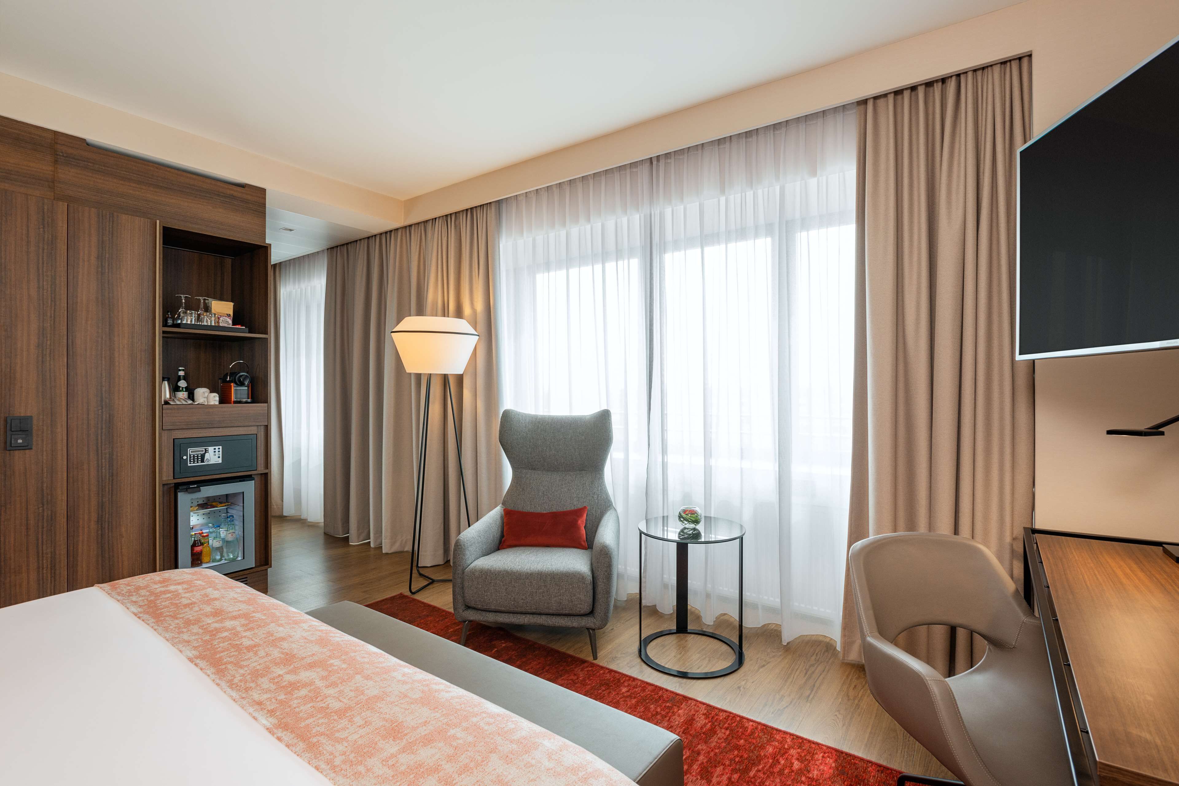A hotel room with a bed, a desk, a chair, a small table, a lamp, and a minibar.