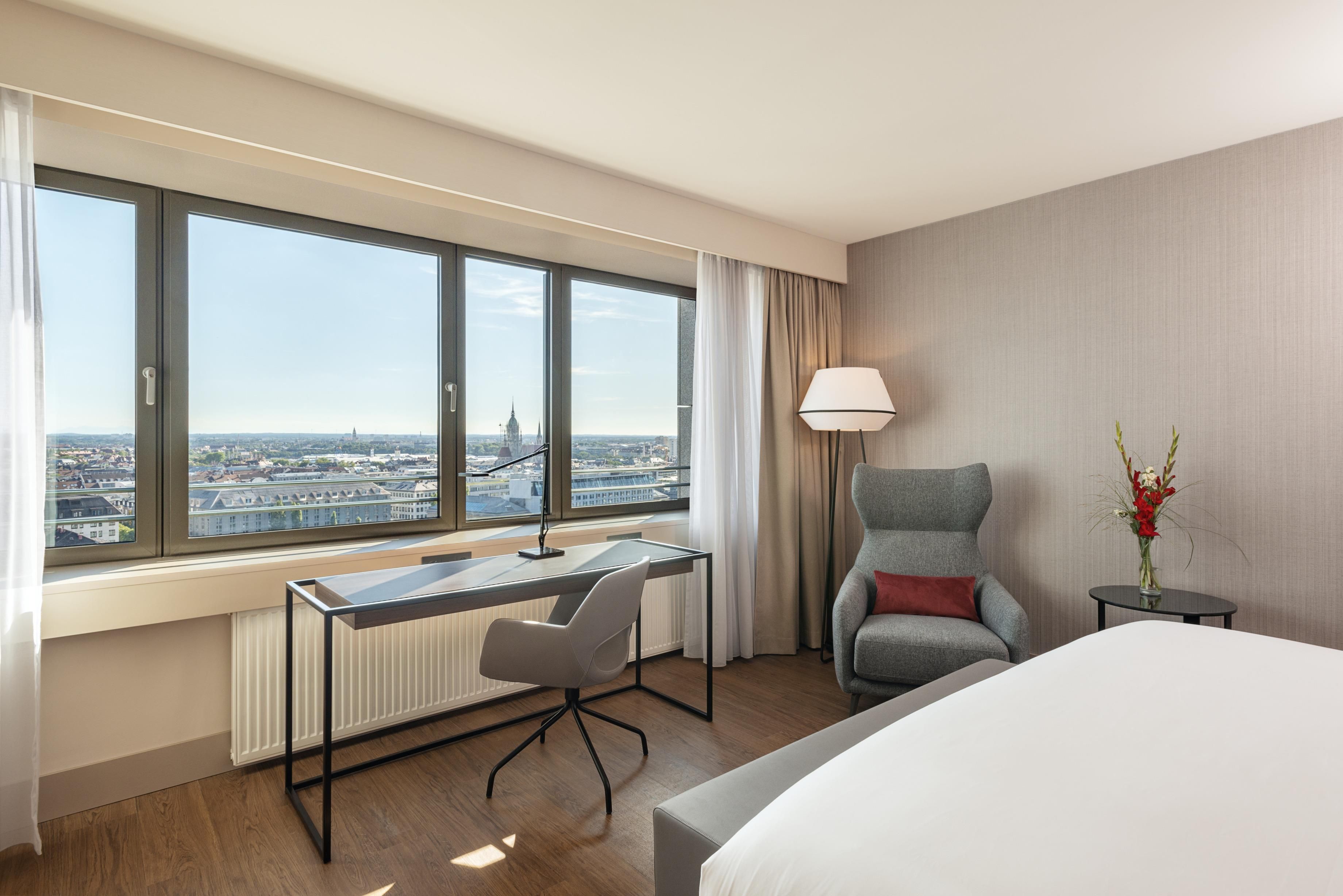 Habitación de hotel con un ventanal grande, un escritorio, una silla, una cama y una pequeña mesa con un jarrón de flores.
