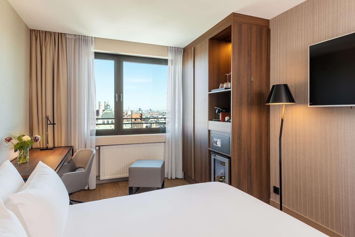 A hotel room with a large bed, a desk with a chair, a small table with flowers, a radiator, a TV mounted on the wall, and a view of the city through the window.