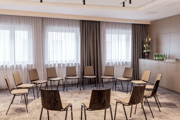 A conference room with beige chairs arranged in a semi-circle, large windows with sheer curtains, and a cabinet with bottled beverages.