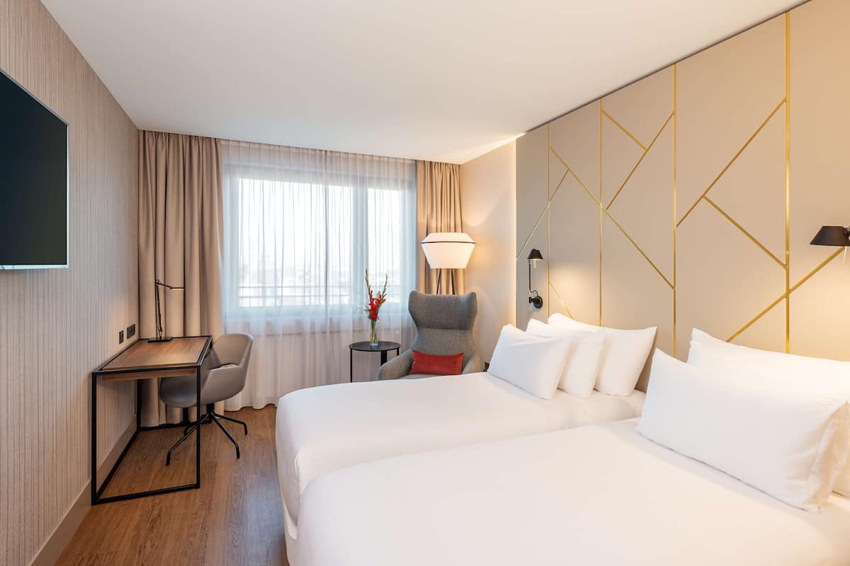 A hotel room with two beds, a desk, a chair, a TV, and a lamp.