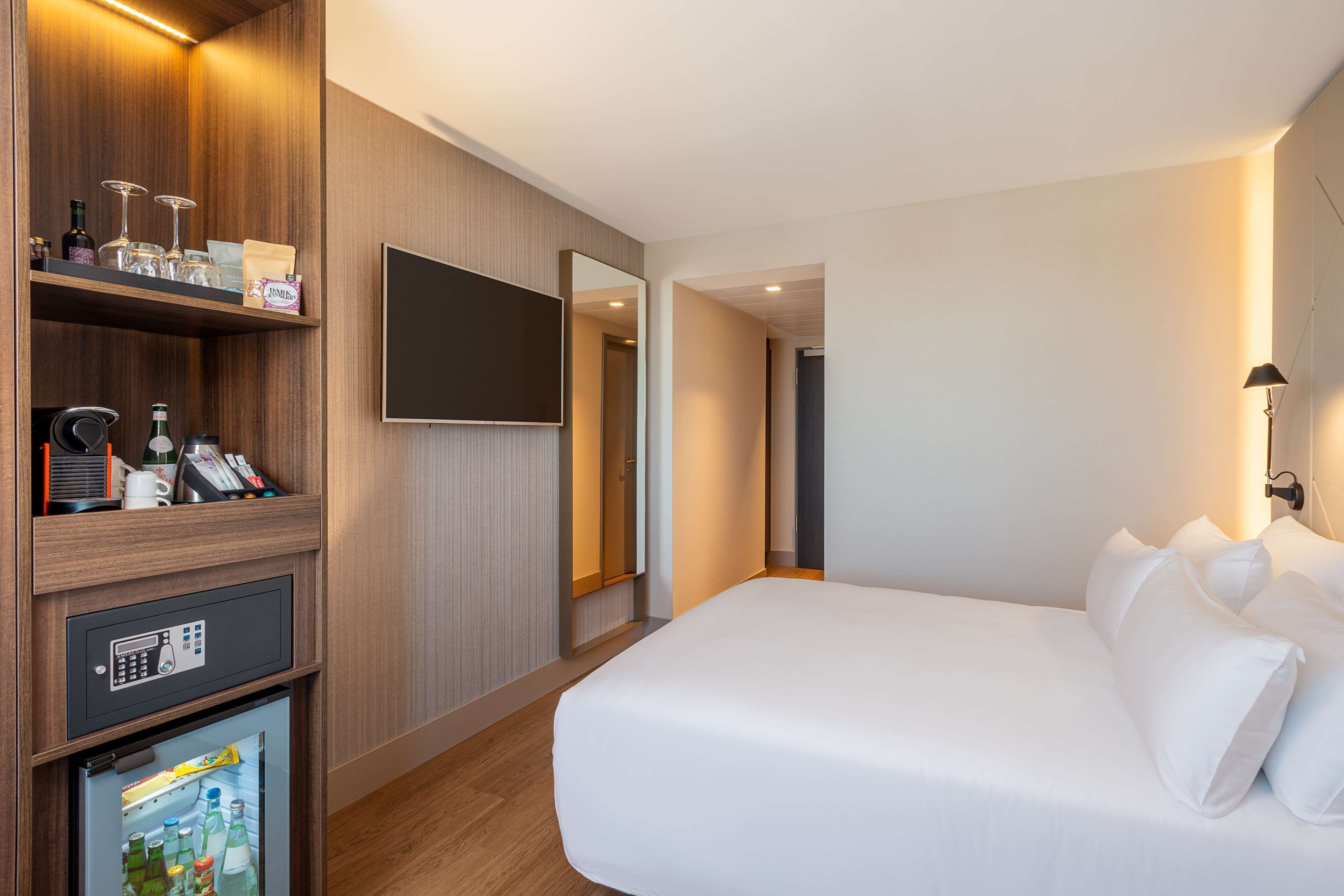 A hotel room with a bed, a TV mounted on the wall, a minibar, and a mirror.