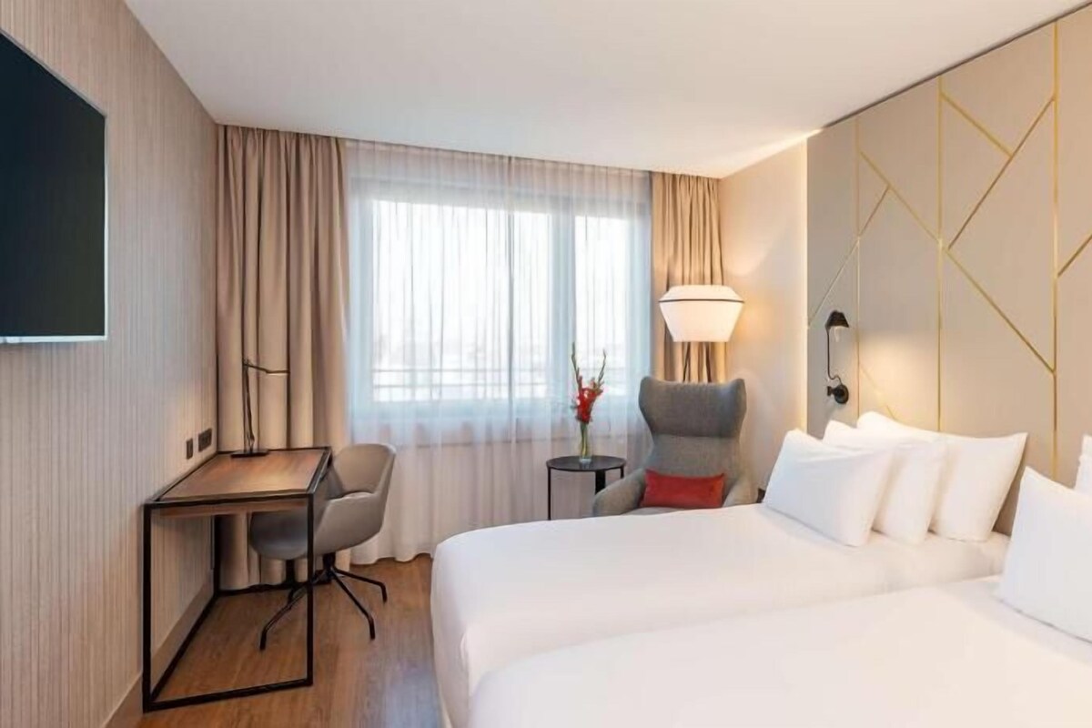 Una camera d'albergo con due letti, una scrivania, una sedia, una lampada e un televisore appeso al muro.