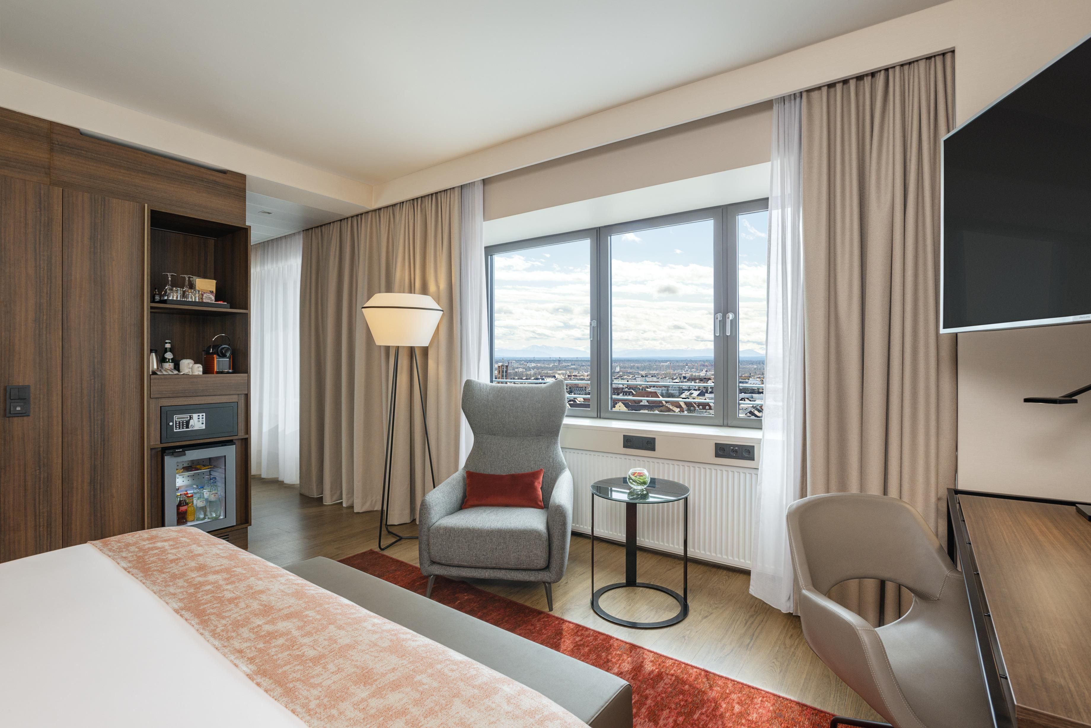 A hotel room with a bed, a desk, a chair, a small table, a lamp, and a large window with a city view.