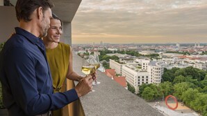 Una pareja sosteniendo copas de vino en un balcón con vistas a un paisaje urbano.