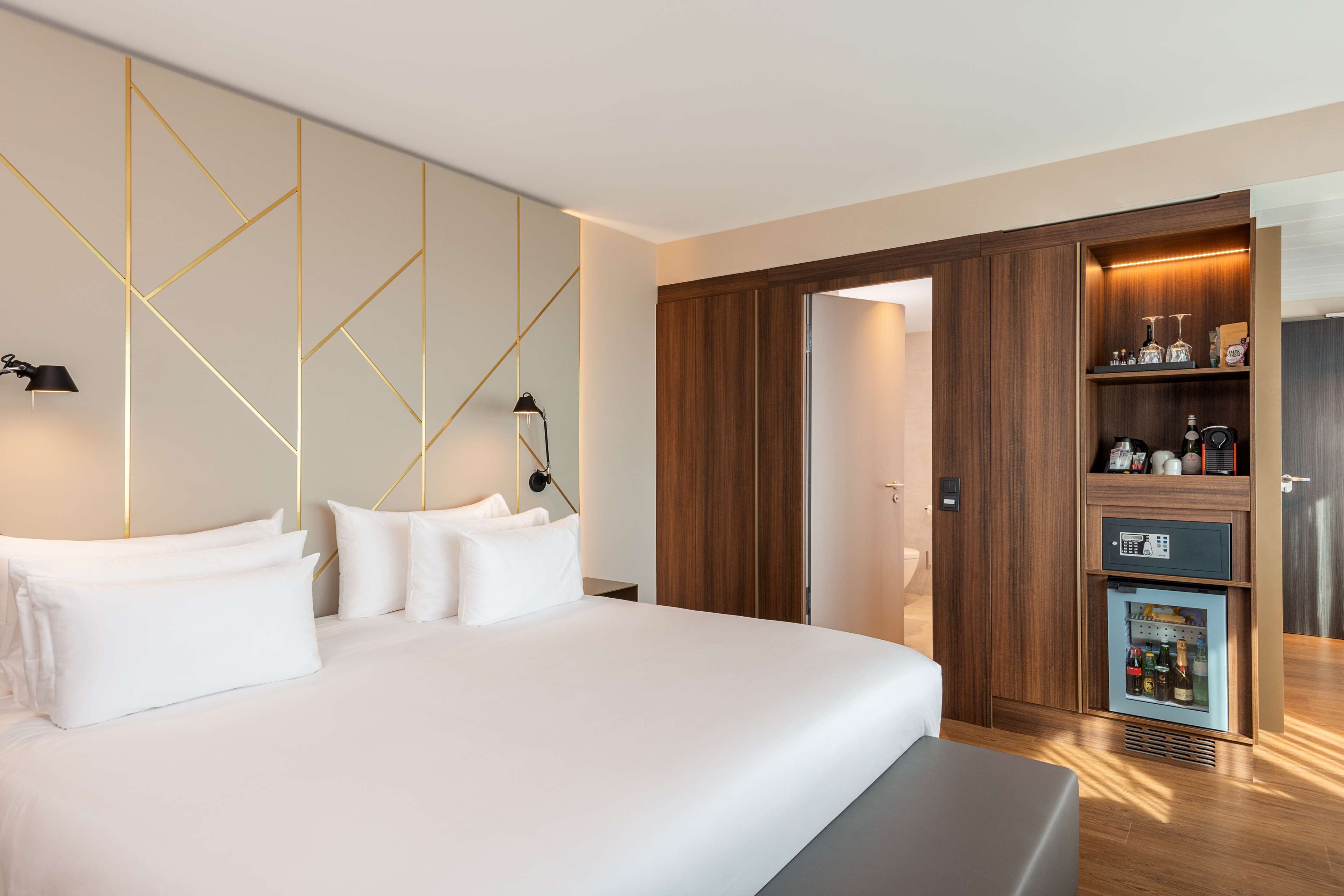 A modern hotel room with a large bed, a built-in wardrobe, and a minibar.