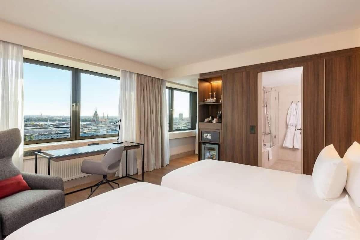 A hotel room with a large window, a desk, two beds, a wardrobe, and a shower area.