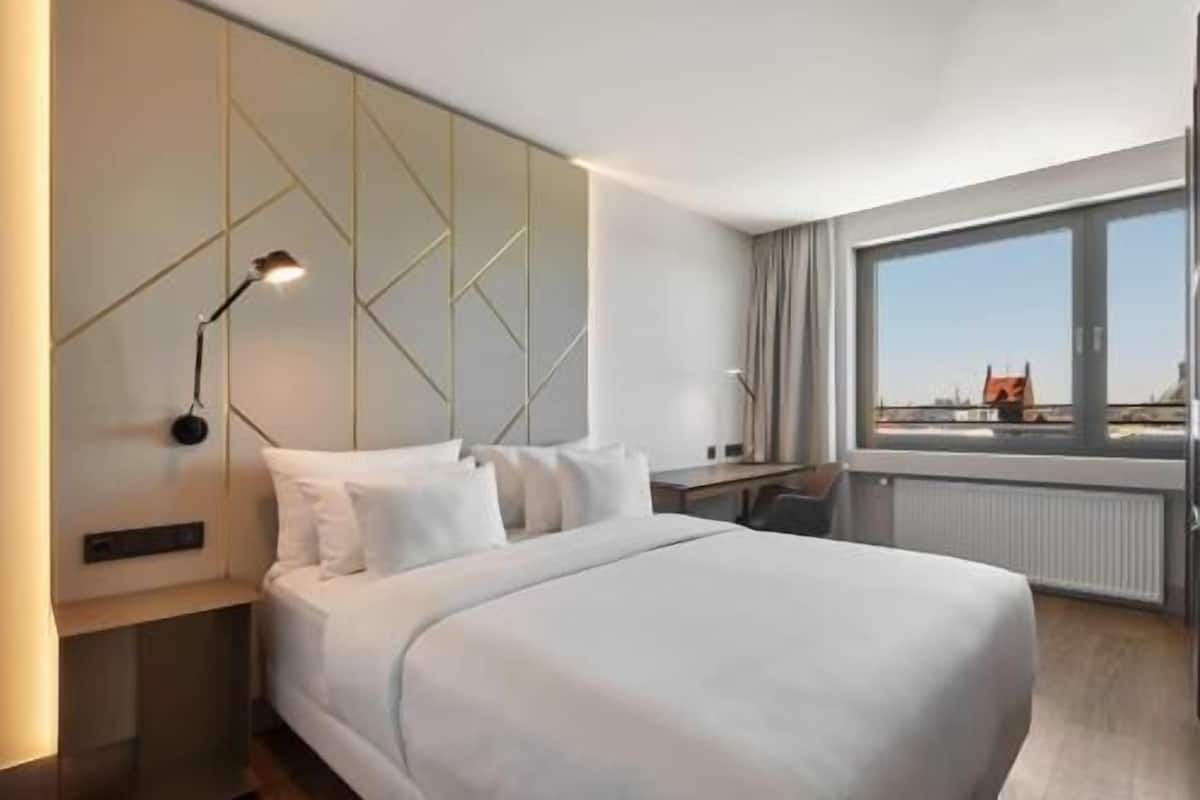 A modern hotel room with a large bed, a desk, and a view of the city.