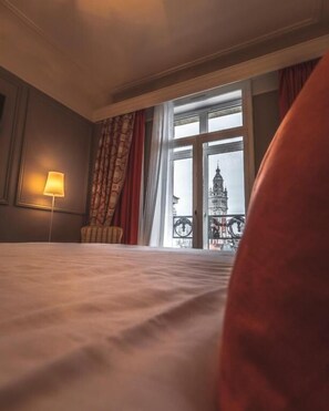 Habitación de hotel con cama, lámpara, ventana con cortinas y vista a un edificio.