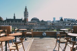 Una terraza en la azotea con mesas y sillas de madera, que ofrece vistas a edificios históricos y rascacielos modernos.