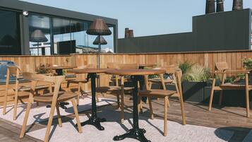 An outdoor seating area with wooden tables and chairs, a lamp, and a view of a modern building with large windows and chimneys.