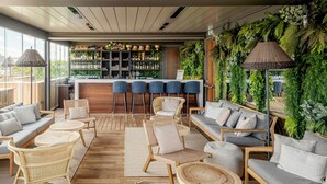 A modern bar area with a wooden bar counter, multiple seating options including bar stools and cushioned chairs, and a backdrop of lush green plants.